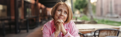 An older woman sits outdoors, deep in thought, with her hands folded