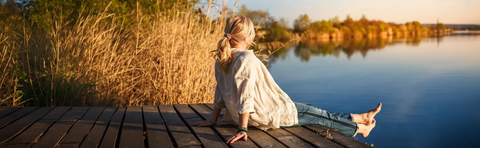 A woman is sitting on a wooden boardwalk by the lake in the evening light, with reeds in the foreground