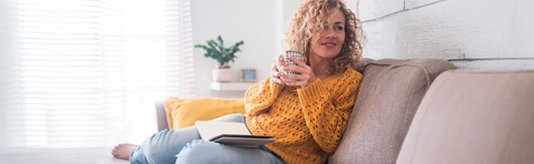 Frau mit Locken sitzt mit Tasse und Buch auf dem Sofa