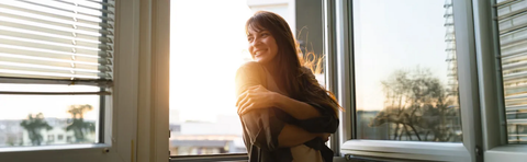 A woman is sitting by the window, smiling in the backlight of the low-hanging sun, with her arms wrapped around her knees
