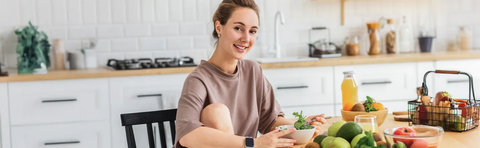 A woman is sitting at a table in a kitchen with fresh fruits and vegetables