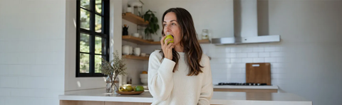 A woman is eating an apple in a bright kitchen; Image note: AI-assisted content