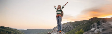 A young woman with a backpack is standing on a rock and raising her arms high above the mountains at sunset.