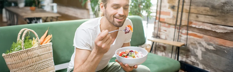 Mann isst eine Schale Müsli auf einem grünen Sofa, daneben ein Gemüsekorb