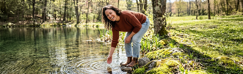 Eine lächelnde Frau in Jeans und braunem Pullover beugt sich am Ufer eines idyllischen Waldsees vor und berührt mit der Hand das klare Wasser; Bildhinweis: KI-gestützter Inhalt