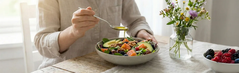 A person is eating a salad at a table, with flowers and a bowl of berries next to them; Image note: AI-assisted content