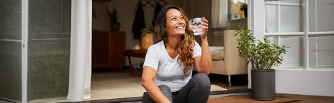 A smiling woman is sitting on the floor of a bright living room, drinking from a glass
