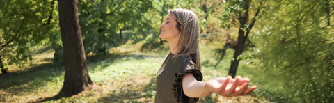 A woman stands with her arms outstretched and her eyes closed in a sunny, green park