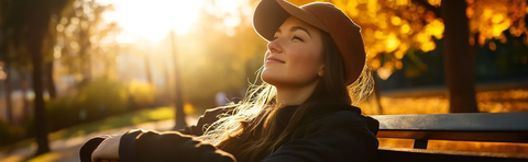 Frau mit Hut sitzt auf einer Bank im Herbstlaub, das Gesicht zur Sonne
