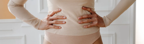 A woman wearing a beige top places both hands on her sides near her stomach; a bright room in the background