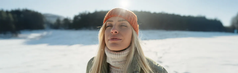 A woman wearing a hat is standing in the snow, her face turned toward the sun
