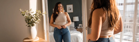 A woman in jeans and a light-colored top is standing in front of a bed, looking at her stomach.; Image note: AI-assisted content