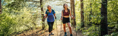 Two smiling women are hiking through a sun-drenched forest with hiking poles