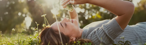 A smiling woman is lying in a sunny flower meadow and holding a flower above her head