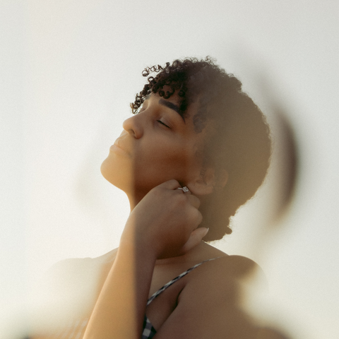 A woman with curly hair and her eyes closed, her hand on her neck, bathed in warm backlighting