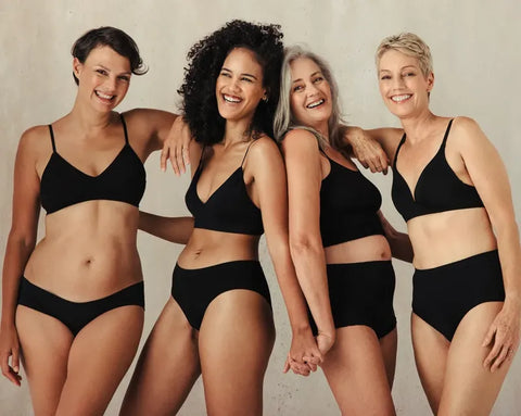 Four smiling women of different ages, wearing black underwear, are standing next to each other