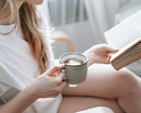 A woman holding a cup of tea in one hand and an open book in the other