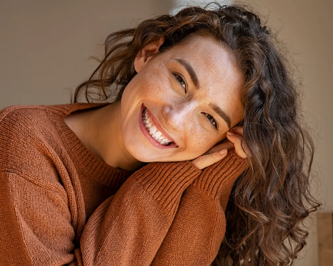 A smiling woman with curly hair and freckles, resting her head on her hand