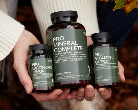 Zwei Hände halten Pro Lactoferrin Immun, Pro Mineral Complete und Pro Vitamin C Ultra vor herbstlichem Laub