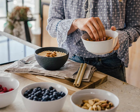 Eine Person in einer hellen Küche fügt Nüsse aus einer Schale zu ihrem Frühstücks-Porridge hinzu; im Vordergrund stehen frische Beeren