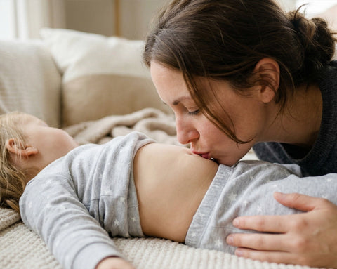 Eine Mutter beugt sich über ihr schlafendes Kleinkind auf dem Sofa und gibt ihm einen sanften Kuss auf den Bauch; Bildhinweis: KI-gestützter Inhalt