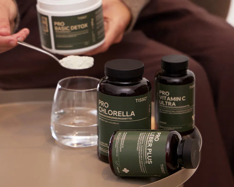 Das Detox-Paket von TISSO mit Pro Leber Plus, Pro Chlorella und Pro Vitamin C Ultra, die neben einem Glas Wasser auf einem Tisch stehen, während Pro Basic Detox in der Hand gehalten wird und daraus ein  Löffel Pulver über dem Glas schwebt