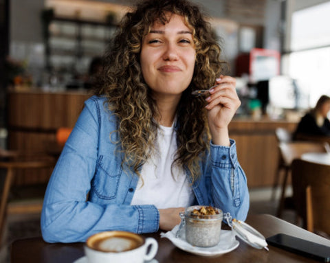 Eine lächelnde Frau mit lockigem Haar sitzt in einem Café an einem Tisch und isst mit einem Löffel ein Dessert aus einem Glas, neben einer Tasse Cappuccino.