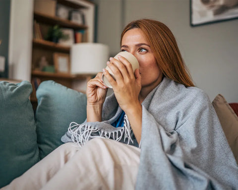 Eine Frau mit langen rötlich-braunen Haaren sitzt eingekuschelt in eine graue Decke auf dem Sofa und trinkt aus einer Tasse.
