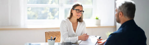 A therapist wearing glasses is conducting a consultation at a desk; across from her is a client with documents.